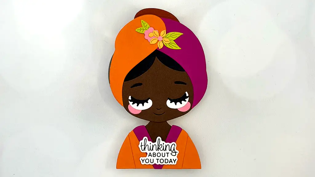 A handmade card features a smiling woman with brown skin, an orange and purple head wrap, and a sign reading "thinking about you today." Perfect for card making enthusiasts and fans of Stunning Cards from the Spellbinders December Club Kits.