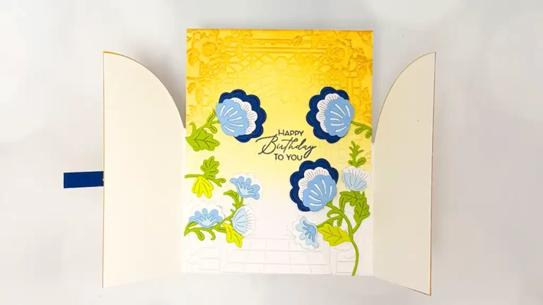 A birthday card with embossed yellow top, blue and green floral designs, and the text “Happy Birthday to You” in the center. Perfect for cardmaking enthusiasts, this Regal Reflections card is shown partially open.