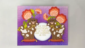 Greeting card with two brown ceramic dog figurines holding orange flowers, purple gradient background, and the message: "May your birthday be as timeless and charming as you.