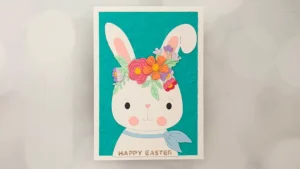 Illustrated Easter card featuring a white bunny with a blue scarf and a colorful flower crown, set against a teal background with "Happy Easter" text at the bottom. Created with Spellbinders Feb 26 Club kits